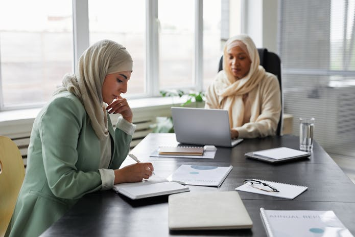 Two women in hijabs seated at a table, working on Razer Blade 17 laptops, engaged in focused discussion.